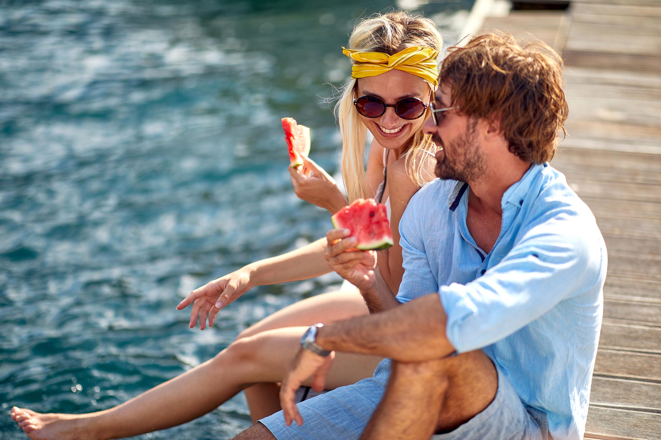 Close up shot of couple with watermelon enjoying sunny day on wooden jetty by water. Tourism, summertime, togetherness, lifestyle concept.